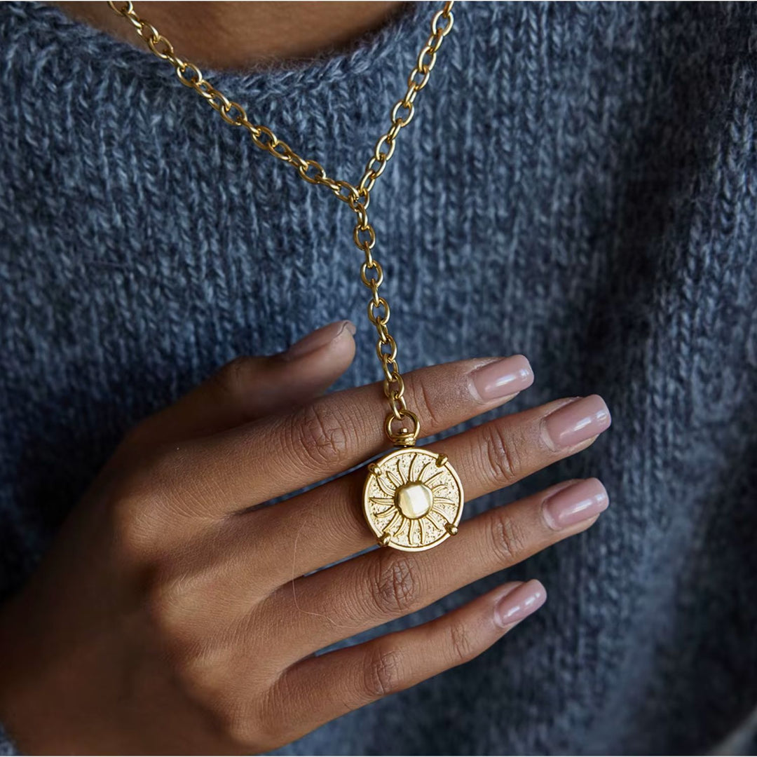 Gold necklace with a circular pendant held by a hand against a textured gray background