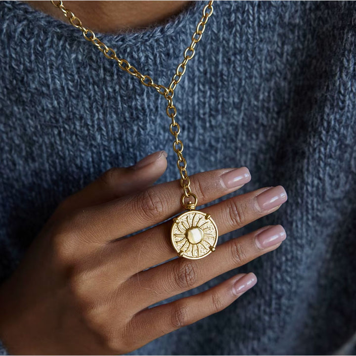 Gold necklace with a circular pendant held by a hand against a textured gray background
