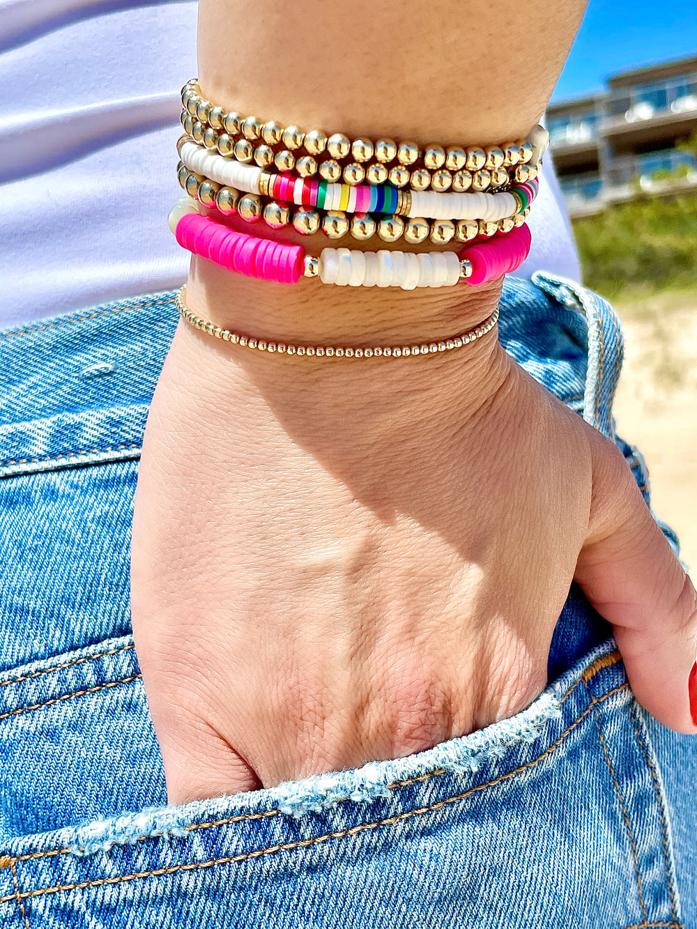 Olivia le model wearing the rainbow seychelles bead bracelet on her wrist layered with the nirvana hot pink pearl bracelet and 4mm gold filled bubble bead bracelet all layered together and her hand is in her back pocket of her jeans showing off a cool look.