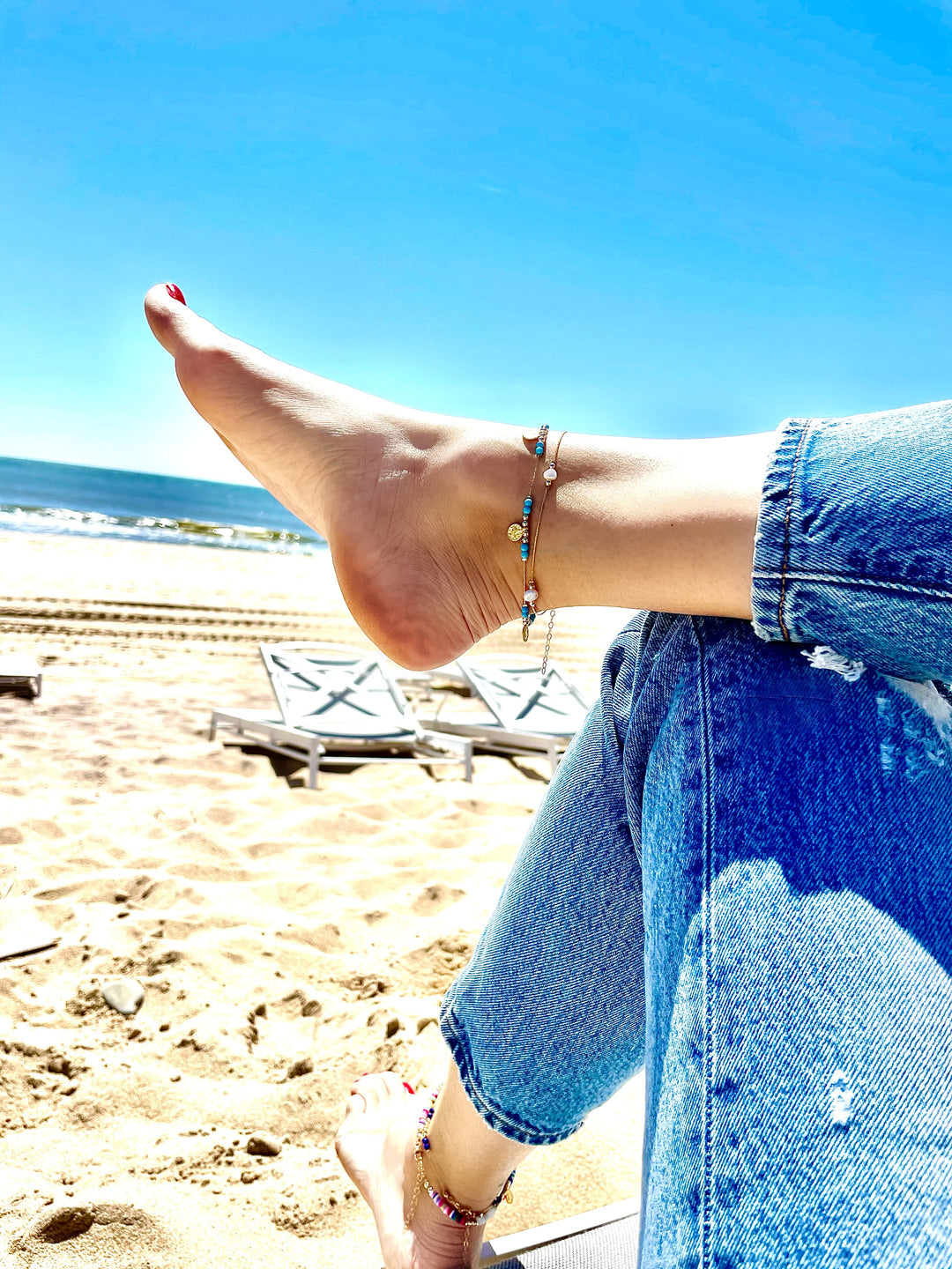 Olivia le model wearing journey pearl anklet layered with journey turq magnesite anklet on the beach wearing jeans and crossing leg over to show the anklets